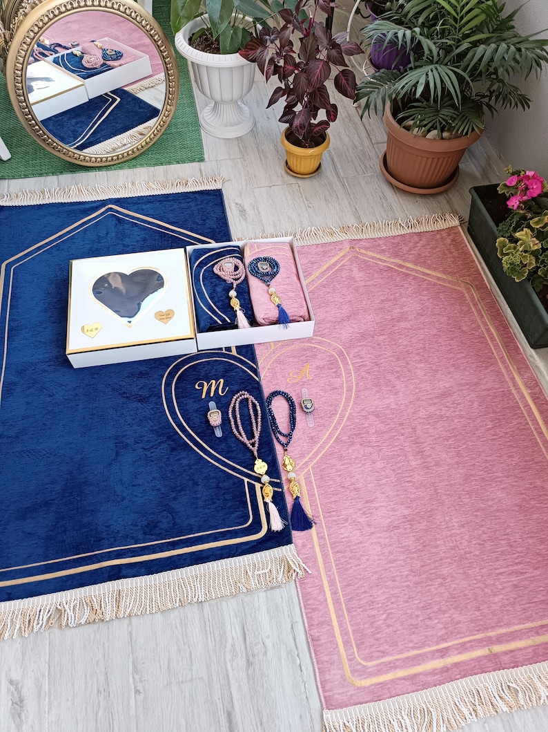 May include: Two prayer rugs, one blue and one pink, with gold trim and tassels. Each rug has a heart-shaped design with a gold outline. A small white box with a heart-shaped cutout is in the center of the image. The box contains two sets of prayer beads.