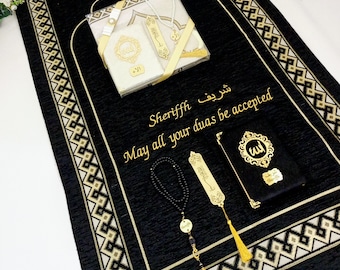 May include: A black prayer rug with gold and beige geometric patterns. The rug is decorated with a gold tassel, a gold bookmark, a gold prayer bead necklace, and a small gold prayer book. The rug is personalised with the name "Sherif" and the text "May all your duas be accepted".