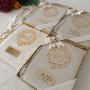 K&ouml;nnte beinhalten: Ein Set rechteckiger Geschenkboxen mit beigem Samtbezug, jeweils mit einem goldenen Filigran-Emblem mit arabischer Kalligrafie, einer wei&szlig;en Schleife mit Perlenakzent, einer wei&szlig;en Perlenkette und einem personalisierten goldenen Namensschild.
