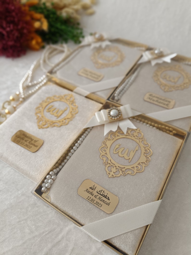 K&ouml;nnte beinhalten: Ein Set rechteckiger Geschenkboxen mit beigem Samtbezug, jeweils mit einem goldenen Filigran-Emblem mit arabischer Kalligrafie, einer wei&szlig;en Schleife mit Perlenakzent, einer wei&szlig;en Perlenkette und einem personalisierten goldenen Namensschild.