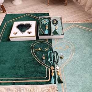 May include: Two green prayer rugs with gold trim and tassels. One rug has a white box with a heart-shaped window and a set of prayer beads. The other rug has a set of prayer beads and a digital tasbih.