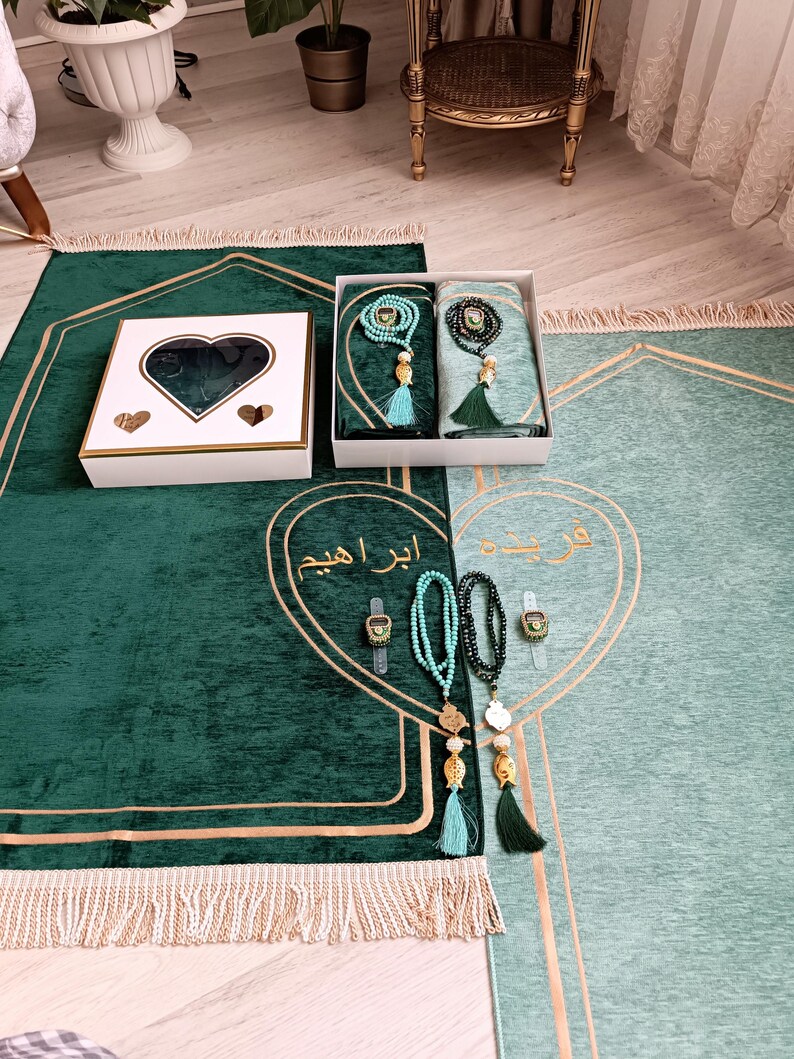 May include: Two green prayer rugs with gold trim and tassels. One rug has a white box with a heart-shaped window and a set of prayer beads. The other rug has a set of prayer beads and a digital tasbih.