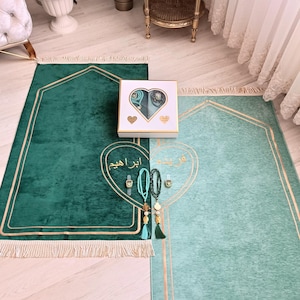 May include: Two green prayer rugs with gold trim and fringe. The rugs are laid out on a wooden floor. A white box with a heart-shaped design contains prayer beads and other religious items. The box is labeled "Abrar Aqeem".