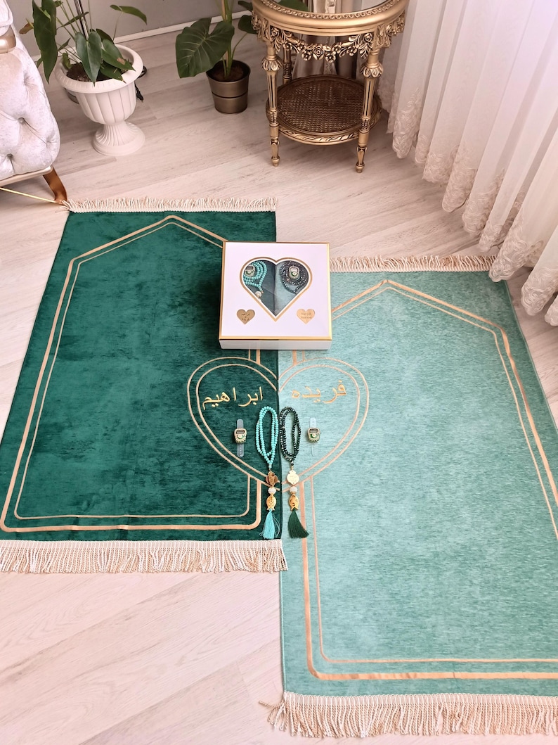 May include: Two green prayer rugs with gold trim and fringe. The rugs are laid out on a wooden floor. A white box with a heart-shaped design contains prayer beads and other religious items. The box is labeled "Abrar Aqeem".