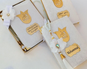 May include: Three white fabric-covered books with gold-toned metal accents, including a tulip-shaped emblem with Arabic calligraphy, a nameplate reading Sarah Alcheikh or Mohammad, and a white beaded prayer bead strand with a white flower tassel.