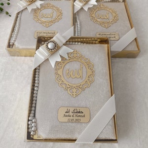 K&ouml;nnte beinhalten: Drei rechteckige Geschenkboxen mit einem Gebetbuch mit beigem Samteinband, einem goldfarbenen Metallemblem mit arabischer Kalligrafie und einer Kette aus wei&szlig;en Gebetsperlen. Jede Box ist mit einem wei&szlig;en Band und einer Strassbrosche verziert.