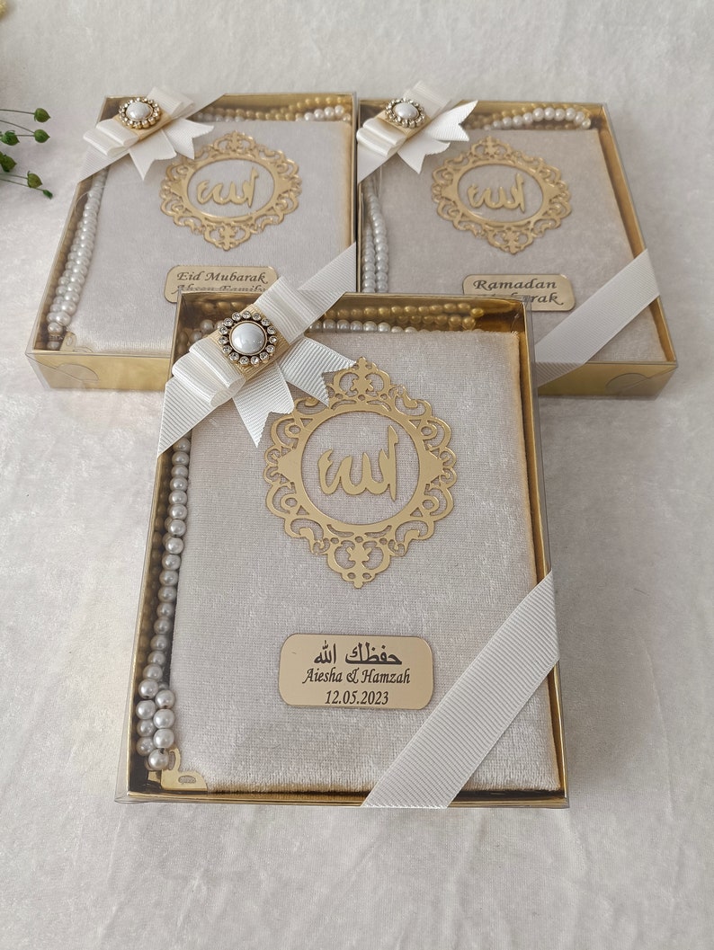 K&ouml;nnte beinhalten: Drei rechteckige Geschenkboxen mit einem Gebetbuch mit beigem Samteinband, einem goldfarbenen Metallemblem mit arabischer Kalligrafie und einer Kette aus wei&szlig;en Gebetsperlen. Jede Box ist mit einem wei&szlig;en Band und einer Strassbrosche verziert.