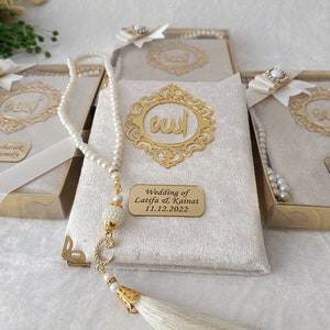 K&ouml;nnte beinhalten: Ein Set rechteckiger Geschenkboxen mit cremefarbenem Samtbezug, jeweils mit einem goldfarbenen verzierten Rahmen mit arabischer Kalligrafie, einer wei&szlig;en Schleife mit Perlenakzent und einer wei&szlig;en Gebetskette mit goldener Quaste.