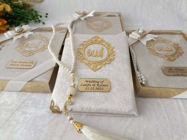 K&ouml;nnte beinhalten: Ein Set rechteckiger Geschenkboxen mit cremefarbenem Samtbezug, jeweils mit einem goldfarbenen verzierten Rahmen mit arabischer Kalligrafie, einer wei&szlig;en Schleife mit Perlenakzent und einer wei&szlig;en Gebetskette mit goldener Quaste.
