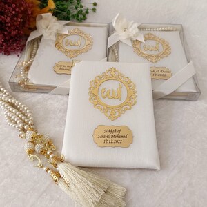 Pu&ograve; includere: Tre libri rettangolari bianchi con cornici in filigrana dorata contenenti calligrafia araba, ciascuno decorato con un fiocco di nastro bianco. Una lunga collana di perle bianche con accenti dorati e una nappa riposa accanto ai libri.