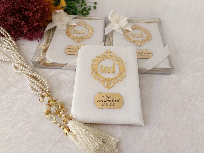 Pu&ograve; includere: Tre libri rettangolari bianchi con cornici in filigrana dorata contenenti calligrafia araba, ciascuno decorato con un fiocco di nastro bianco. Una lunga collana di perle bianche con accenti dorati e una nappa riposa accanto ai libri.