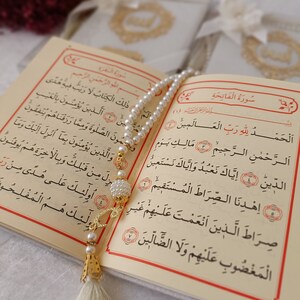 Pu&ograve; includere: Un Corano aperto con pagine color crema, testo arabo nero e bordi rossi. Una collana di perle bianche con dettagli dorati e una lunga nappa color crema poggia sulle pagine aperte. Sono visibili due libri coordinati con copertina rigida.