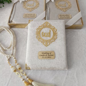 K&ouml;nnte beinhalten: Drei rechteckige Geschenkboxen mit cremefarbenem Samtbezug, jeweils mit einem goldfarbenen, verzierten Rahmen mit arabischer Kalligrafie, einer wei&szlig;en Schleife mit Perlenakzent und einer personalisierten goldenen Plakette mit der Aufschrift Eid Mubarak, Ramadan Mubarak oder Wedding of Latifa & Kainat 11.12.2022, dazu eine lange Kette aus wei&szlig;en Perlen-Gebetsperlen mit goldenen Quasten.