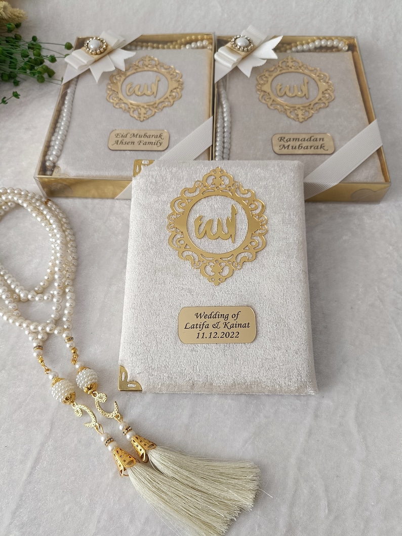K&ouml;nnte beinhalten: Drei rechteckige Geschenkboxen mit cremefarbenem Samtbezug, jeweils mit einem goldfarbenen, verzierten Rahmen mit arabischer Kalligrafie, einer wei&szlig;en Schleife mit Perlenakzent und einer personalisierten goldenen Plakette mit der Aufschrift Eid Mubarak, Ramadan Mubarak oder Wedding of Latifa & Kainat 11.12.2022, dazu eine lange Kette aus wei&szlig;en Perlen-Gebetsperlen mit goldenen Quasten.