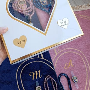 May include: A white heart-shaped box with a gold trim contains two sets of prayer beads, one pink and one blue. The beads are in a heart-shaped compartment with a pink and blue fabric background. The box has a gold heart-shaped tag with the text "Your Gift Note Here". The box is sitting on top of two prayer rugs, one blue and one pink, with gold trim and the letters "A" and "M" embroidered on them.