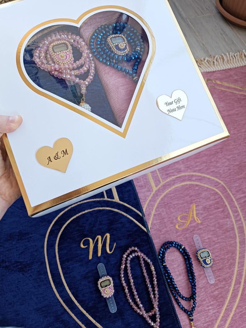 May include: A white heart-shaped box with a gold trim contains two sets of prayer beads, one pink and one blue. The beads are in a heart-shaped compartment with a pink and blue fabric background. The box has a gold heart-shaped tag with the text "Your Gift Note Here". The box is sitting on top of two prayer rugs, one blue and one pink, with gold trim and the letters "A" and "M" embroidered on them.