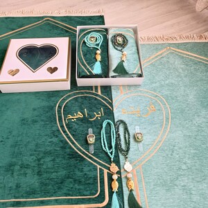 May include: A green prayer rug with gold trim and a white box with a heart-shaped cutout. The rug has a light green section with Arabic text and two sets of prayer beads with tassels. The text on the rug reads "ابراهم".