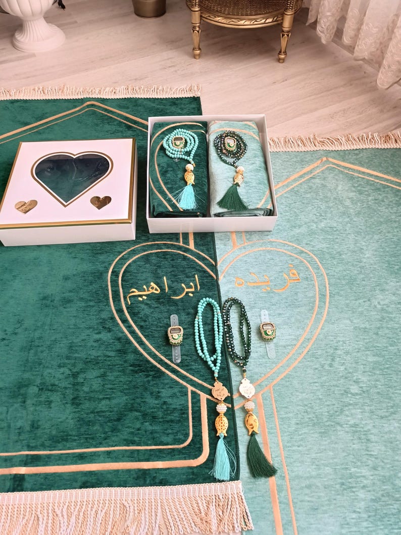 May include: A green prayer rug with gold trim and a white box with a heart-shaped cutout. The rug has a light green section with Arabic text and two sets of prayer beads with tassels. The text on the rug reads "ابراهم".
