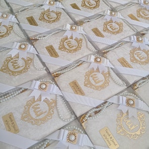 K&ouml;nnte beinhalten: Eine Sammlung quadratischer Geschenkboxen mit wei&szlig;em, strukturiertem Stoff, verziert mit einem goldfarbenen, verzierten Rahmen mit arabischer Kalligrafie, einer wei&szlig;en Schleife mit Perlenakzent und einer Kette aus wei&szlig;en Perlen.