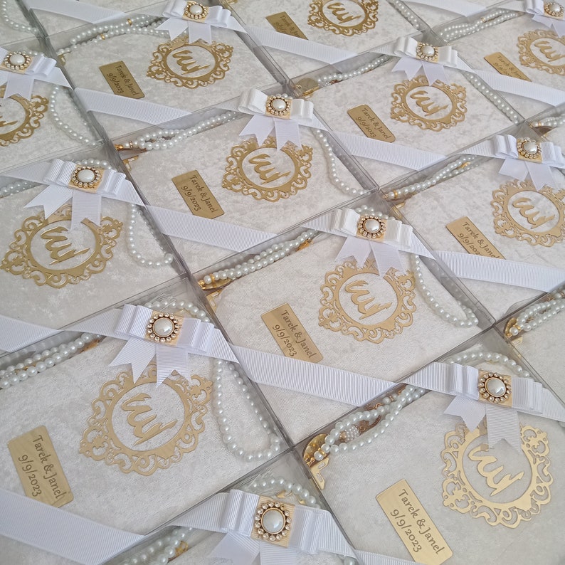 K&ouml;nnte beinhalten: Eine Sammlung quadratischer Geschenkboxen mit wei&szlig;em, strukturiertem Stoff, verziert mit einem goldfarbenen, verzierten Rahmen mit arabischer Kalligrafie, einer wei&szlig;en Schleife mit Perlenakzent und einer Kette aus wei&szlig;en Perlen.