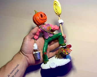 Puede incluir: Una figurita de calabaza pintada a mano con un chaleco morado y pantalones verdes que sostiene un cubo y una piruleta. La figurita está de pie sobre una base con un montón de caramelos de colores.