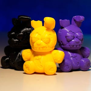 以下が含まれることがあります： 黒、黄色、紫の3Dプリントされたウサギのフィギュアが3つ。 黄色のウサギが中央にあり、左に黒いウサギ、右に紫のウサギがあります。 各ウサギは、詳細で少し脅迫的な表情をしています。