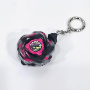 Op de afbeelding: Een zwart en roze tol-sleutelhanger met een centraal rond embleem met een groene draakafbeelding. De tol heeft een bewegingsonscherpte-effect en is bevestigd aan een zilverkleurige metalen ring door een kort zwart plastic verbindingsstuk.