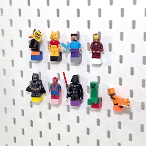 May include: A collection of nine plastic minifigures mounted on a white pegboard. The figures include a black duck holding a sign that reads RABBIT SEASON, a yellow bear, a blocky character with a blue sword, Iron Man, Batman, Spider-Man, Darth Vader with a red lightsaber, a green blocky creature, and an orange fox.