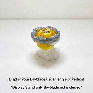 May include: A yellow and grey Beyblade toy on a white display stand. The Beyblade has a yellow centre with a grey outer ring. The display stand is white and has a cross design. Text on the image reads: Display your BeybladeX at an angle or vertical.