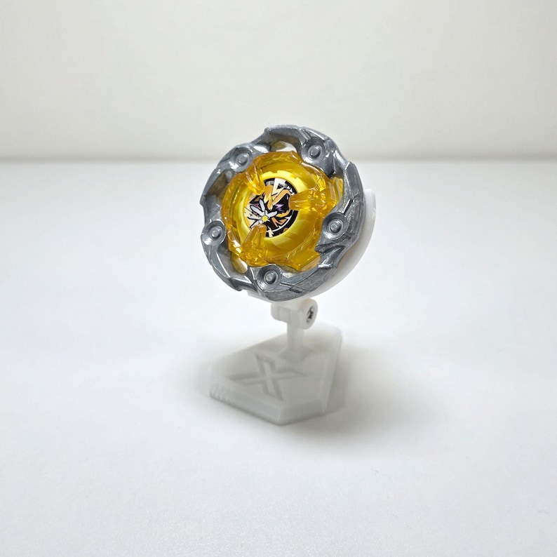 May include: A yellow and silver toy spinning top with a detailed design, mounted on a white stand. The top features a circular shape with intricate patterns and a central design. The stand has a geometric base.