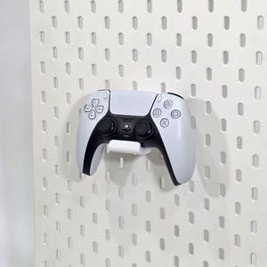 Puede incluir: Un mando blanco y negro de PlayStation 5 montado en una pared perforada blanca. El mando se sujeta con un soporte blanco. La pared perforada tiene una serie de agujeros espaciados.