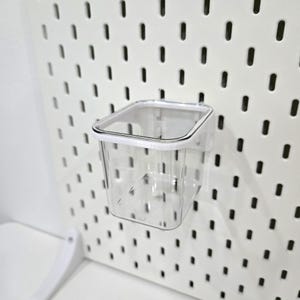 Puede incluir: Un vaso de almacenamiento de plástico transparente, de forma cuadrada, con un borde blanco, diseñado para sujetarse a una panel perforado. El vaso vacío está montado en un panel perforado blanco con agujeros espaciados.