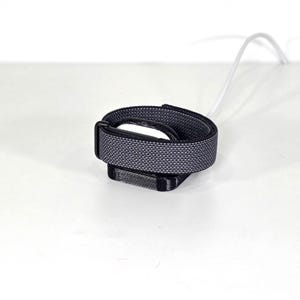 May include: A black and grey woven fabric strap with a rectangular black plastic buckle and a black plastic housing component. A thin white cable extends from the side of the housing, connecting to the strap assembly in a coiled configuration.