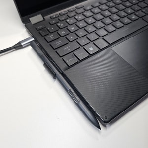 Peut inclure: Un ordinateur portable noir avec un stylet argenté fixé sur le côté. L'ordinateur portable est connecté à un câble noir.