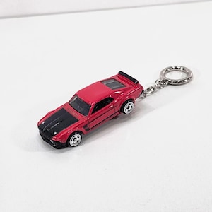 May include: A red and black toy car keychain. The miniature car has a red body with a black hood and a silver keyring. The car has black wheels and a spoiler. The keychain is on a white background.