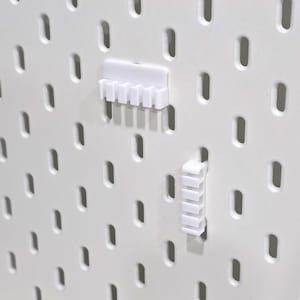 May include: White pegboard with evenly spaced oval holes. Two white plastic pegboard accessories are attached. One accessory has multiple slots, and the other has a series of vertical slots. The pegboard is designed for organization and display.