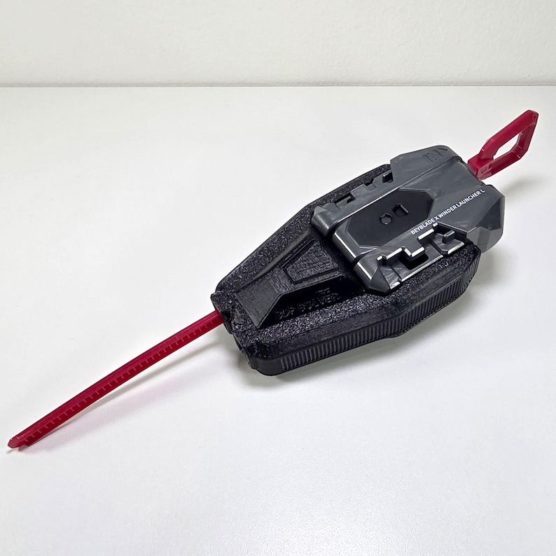 May include: A black and gray Beyblade X Winder Launcher L toy. The launcher has a red pull cord and a red loop. The text "BEYBLADE X WINDER LAUNCHER L" is printed on the top of the launcher.
