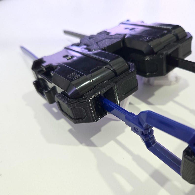 Pu&ograve; includere: Un dispositivo di lancio meccanico in plastica nera caratterizzato da un alloggiamento rettangolare con superfici testurizzate e un meccanismo a strappo in plastica blu che si estende lateralmente. Il dispositivo presenta dettagli stampati.