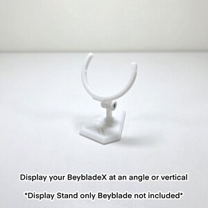 May include: A white display stand for BeybladeX toys. The stand features a curved top and a hexagonal base. The image text says: "Display your BeybladeX at an angle or vertical. *Display Stand only Beyblade not included*"