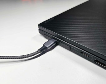 May include: A black laptop with a textured lid and a grey USB-C cable plugged into the side. The laptop has a USB-C port, an HDMI port, and a microSD card slot.