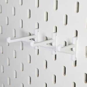 Könnte beinhalten: Zwei weiße Kunststoff-Pegboard-Haken, die an einer weißen Pegboard-Wand montiert sind. Die Haken sind so konzipiert, dass sie Gegenstände am Pegboard halten.