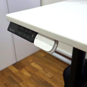 May include: A white desk control panel with two round buttons and the text "S" on the right button. The control panel is attached to the underside of a white desk.