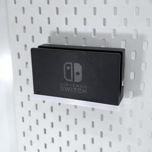 Puede incluir: Un dock negro de Nintendo Switch con el logotipo y el texto de Nintendo Switch. El dock está montado en una placa perforada blanca. Una estantería blanca está debajo.