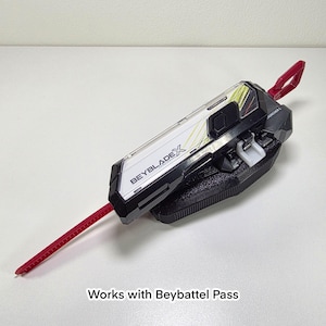 May include: A black, white, and red Beyblade X launcher. The launcher has a red pull cord and a black base. The text "BEYBLADE X" is printed on the side. The text "Works with Beybattel Pass" is printed at the bottom.
