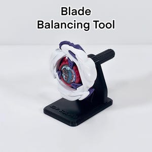 Op de afbeelding: Een zwarte plastic Blade Balancing Tool standaard met de tekst "TOP SOLVER" op de voet. Een wit, paars en rood Beyblade-speeltje is horizontaal op de centrale stang van de standaard gemonteerd, met een cirkelvormig midden met een ontwerp.