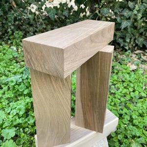 Puede incluir: Una escultura de madera con una parte superior rectangular sostenida por dos columnas verticales. La madera es de color marrón claro natural con vetas visibles. La escultura se exhibe al aire libre sobre un fondo de follaje y hierba verdes.