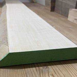 Può includere: Una lunga tavola di legno rettangolare con una superficie superiore liscia e chiara e un bordo grezzo con corteccia naturale su un lato. L'estremità della tavola è tagliata ad angolo e dipinta di un colore verde solido e vivace.