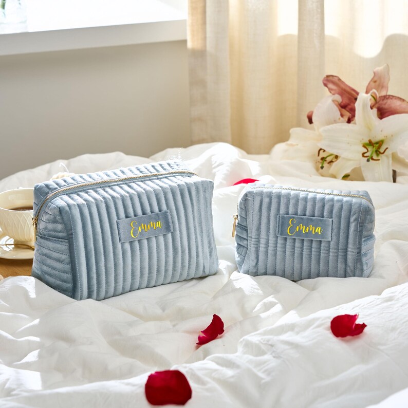 May include: Two light blue, corduroy cosmetic bags with gold zippers and the name "Emma" embroidered in gold. The bags are different sizes and sit on a white bed with red rose petals.