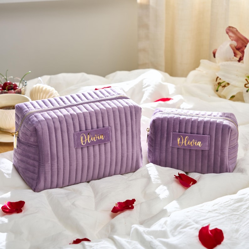 May include: Two lavender-colored, quilted cosmetic bags with gold zippers and the name "Olivia" embroidered in gold. The bags are different sizes and sit on a white bedspread with scattered rose petals.