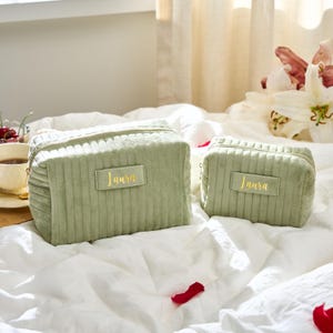May include: Two sage green corduroy cosmetic bags with gold zippers and the name "Laura" in gold lettering. The bags are rectangular and have a quilted design. One bag is larger than the other.